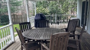 Una terraza de madera cubierta con una mesa redonda y sillas, un ventilador de techo y vistas a los árboles circundantes.