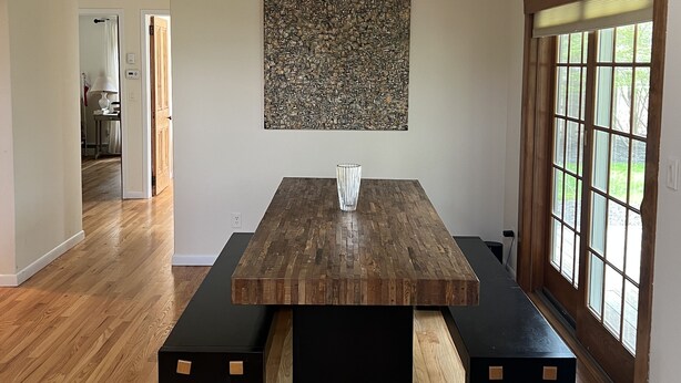 Un comedor con una mesa de madera y bancos, una araña y un ventanal con persianas.