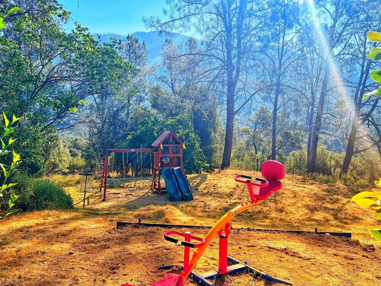 A playground with a slide and swings, surrounded by trees and a clear sky.