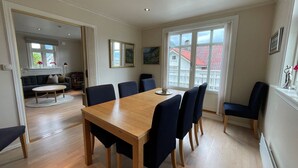 A spacious dining area with a wooden table and blue chairs, a cozy living room with a sofa and a coffee table, and large windows offering a view of a red-roofed building. - Beautiful holiday home in the center of Måløy (Vestland)