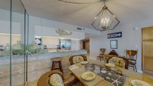 A dining area with wicker chairs, a glass-top table, and a kitchen in the background. - Corner Beachfront. Large Balcony. Pool. Hot Tub. Exceptional Location. (Madeira Beach)