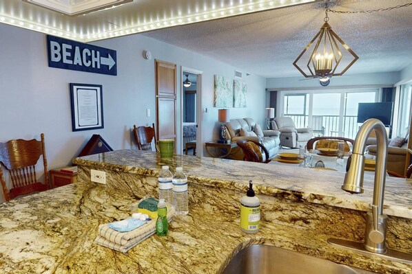 A kitchen with a marble countertop, a sink, and various kitchen items. There is a sign on the wall that says "BEACH" with an arrow pointing to the right.