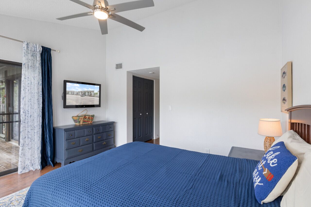 A bedroom with a bed, a television, a dresser, a ceiling fan, and a door.