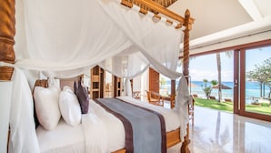 A four-poster bed with white and brown linens, a wooden headboard, and a view of the ocean through large windows. - Villa Dahlia Manggis - 4 bed Oceanfront Retreat, with Chef & Butler Candidasa (Manggis, Karangasem)