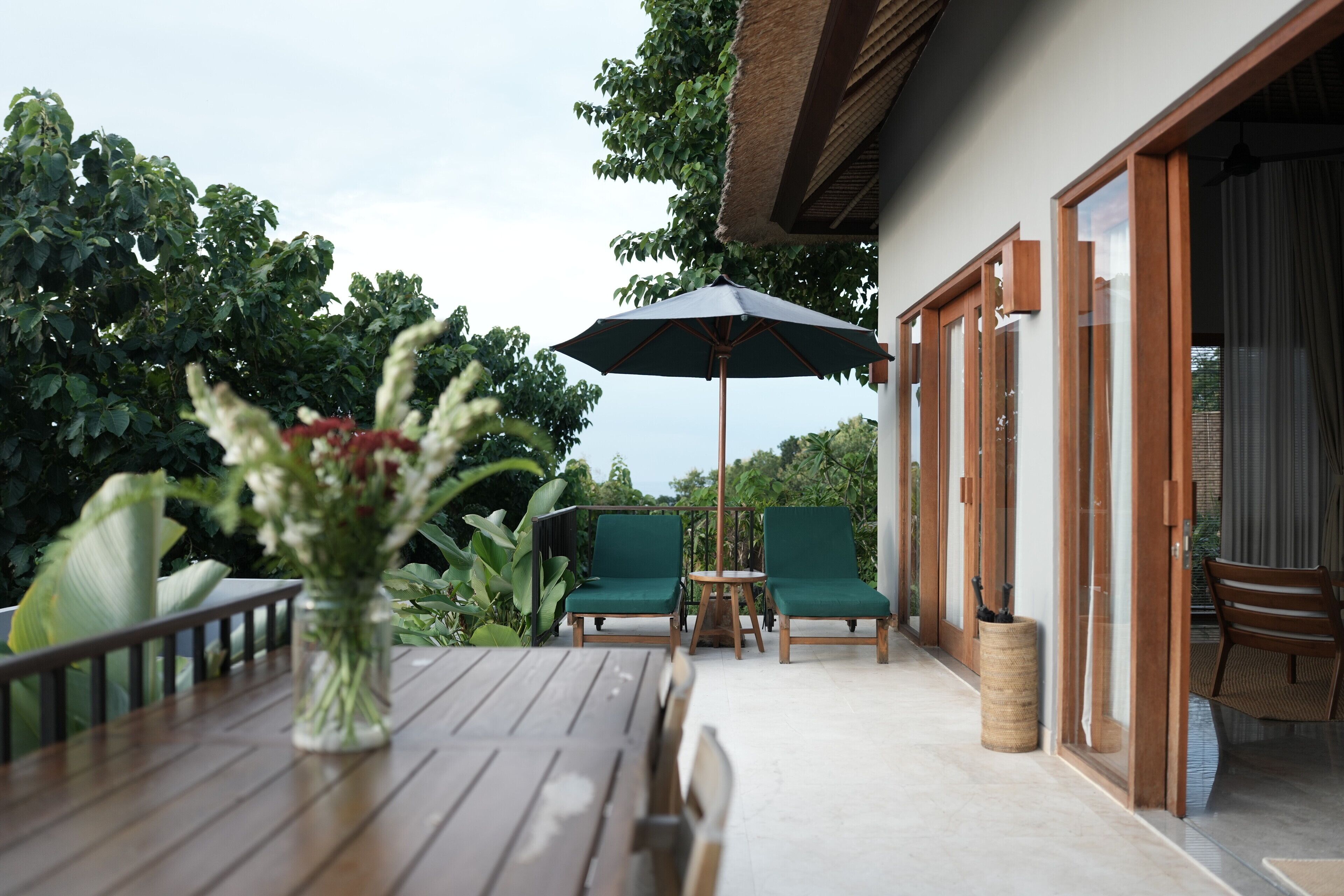 Un espace extérieur aménagé en terrasse, avec une table en bois, deux chaises vertes dotées de coussins, un grand parasol et un vase rempli de fleurs.