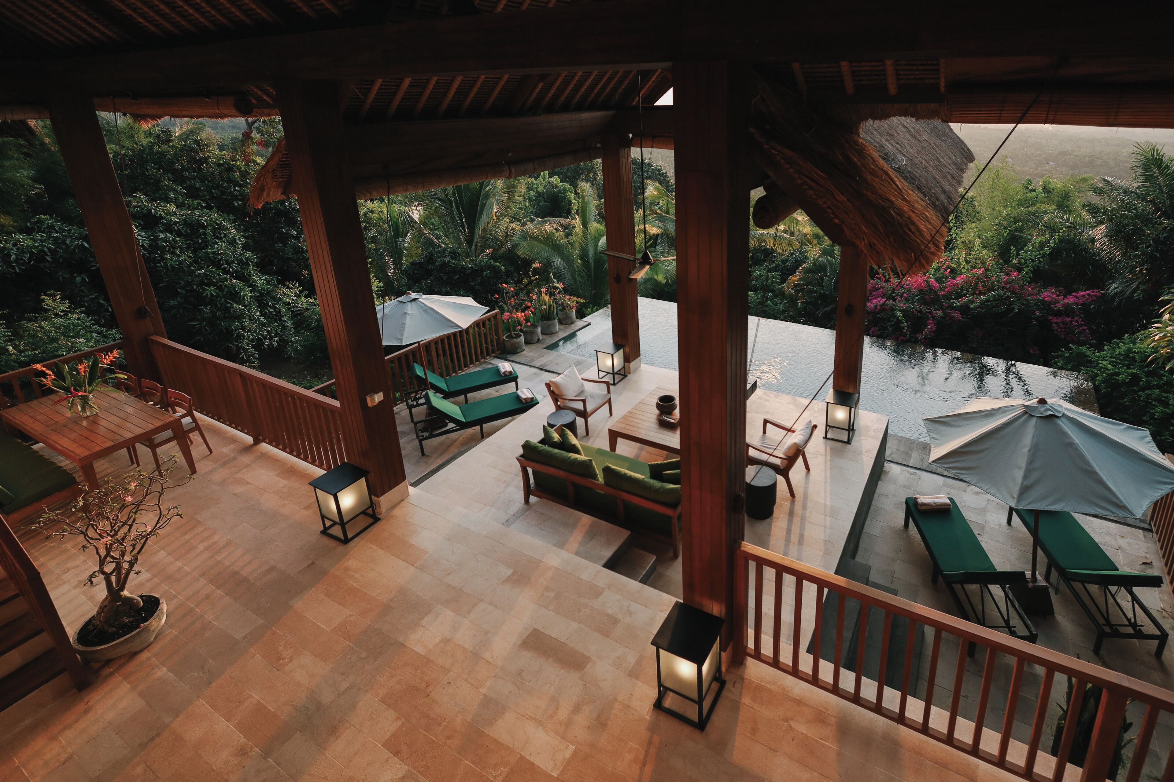A covered outdoor seating area with wooden furniture, a potted plant, and a view of lush greenery and a pool.