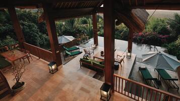 A covered outdoor seating area with wooden furniture, a potted plant, and a view of lush greenery and a pool.