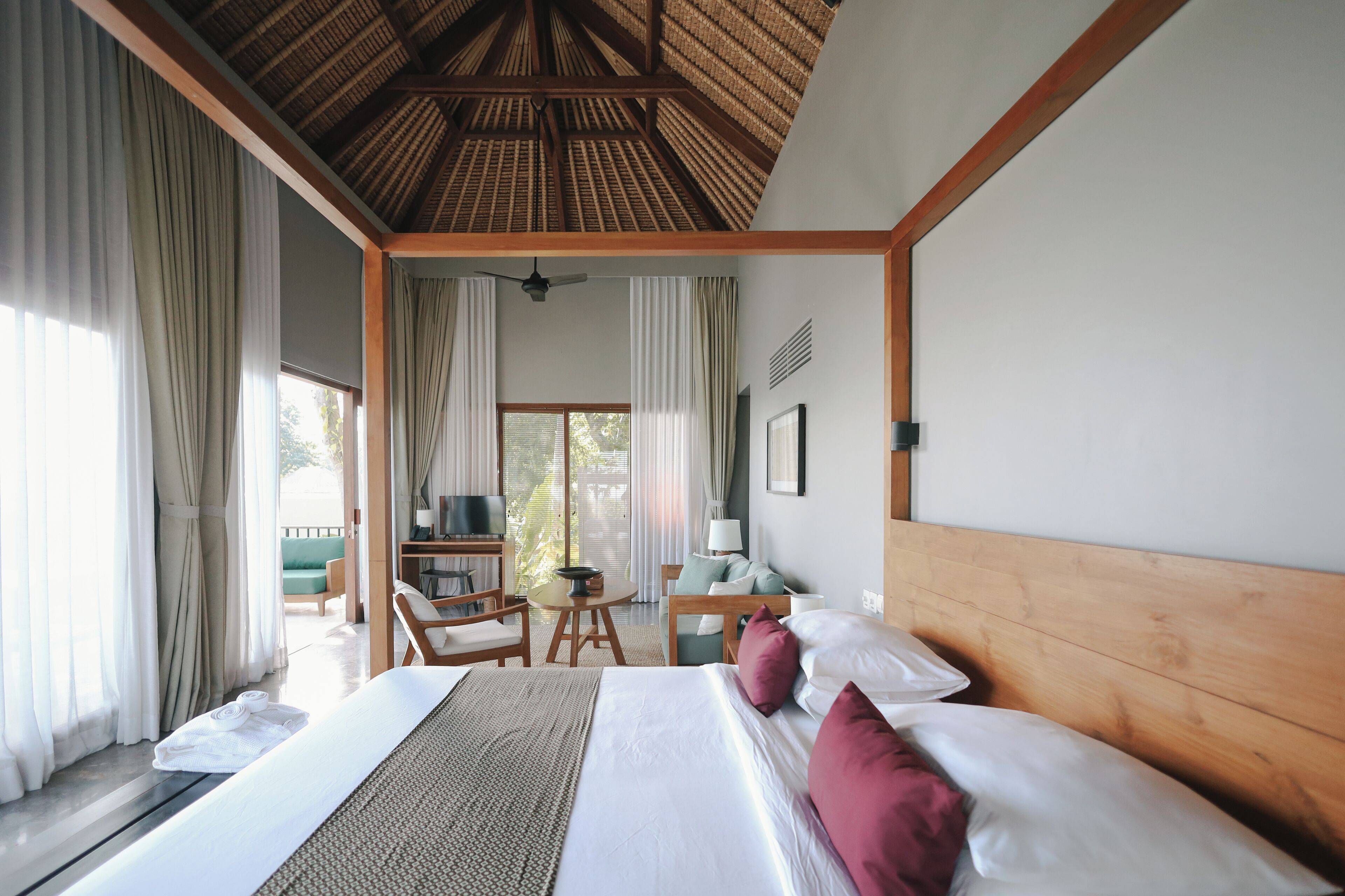 A hotel room with a large bed, a wooden headboard, a desk with a chair, a television, and a balcony with curtains.
