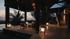 An outdoor patio area with seating and tables, large windows overlooking a body of water, and a staircase leading to an upper level. - Sanglung Villas (Kubutambahan)