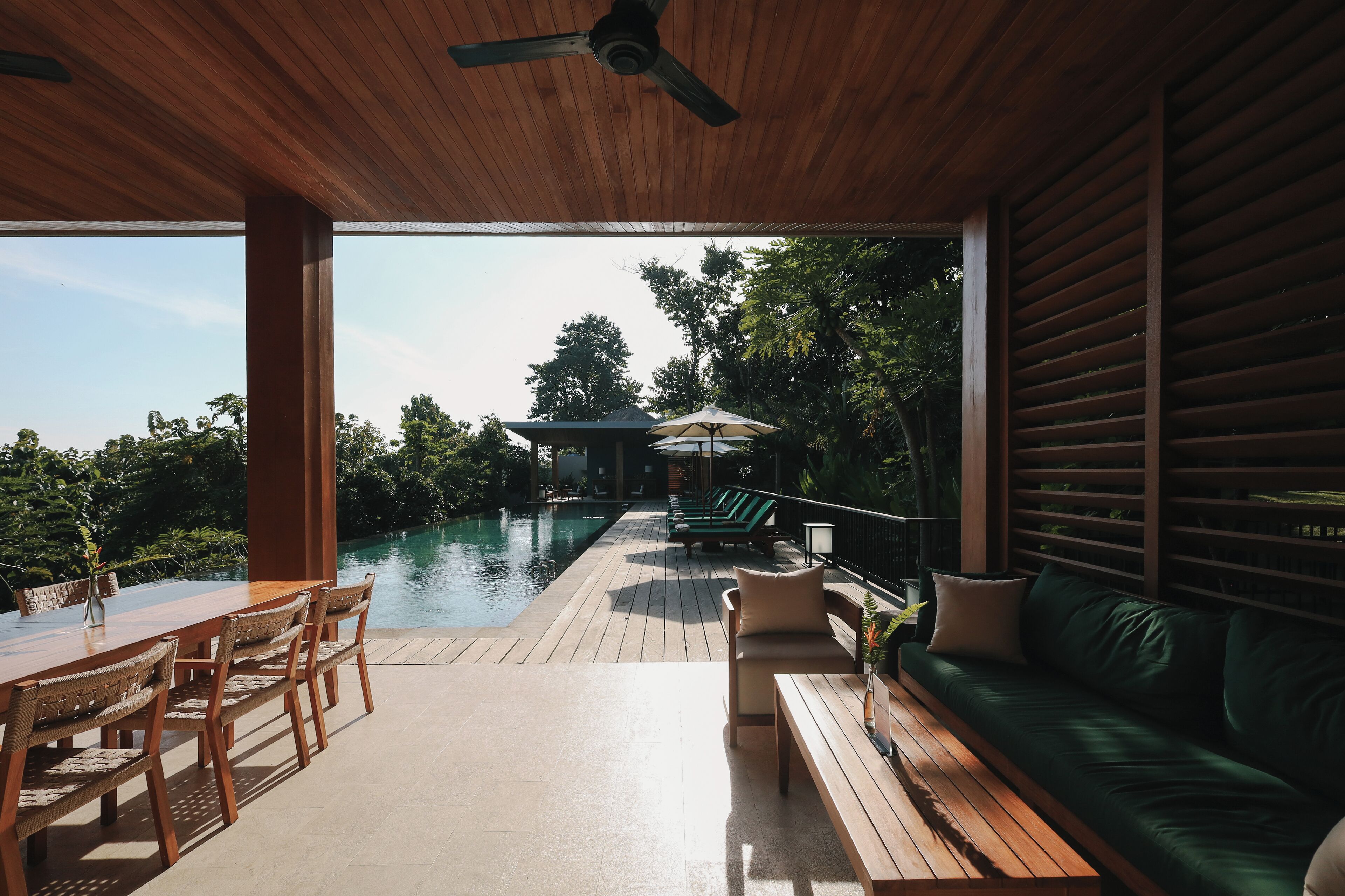 Terraza de madera con piscina, mobiliario exterior y vistas al follaje circundante.