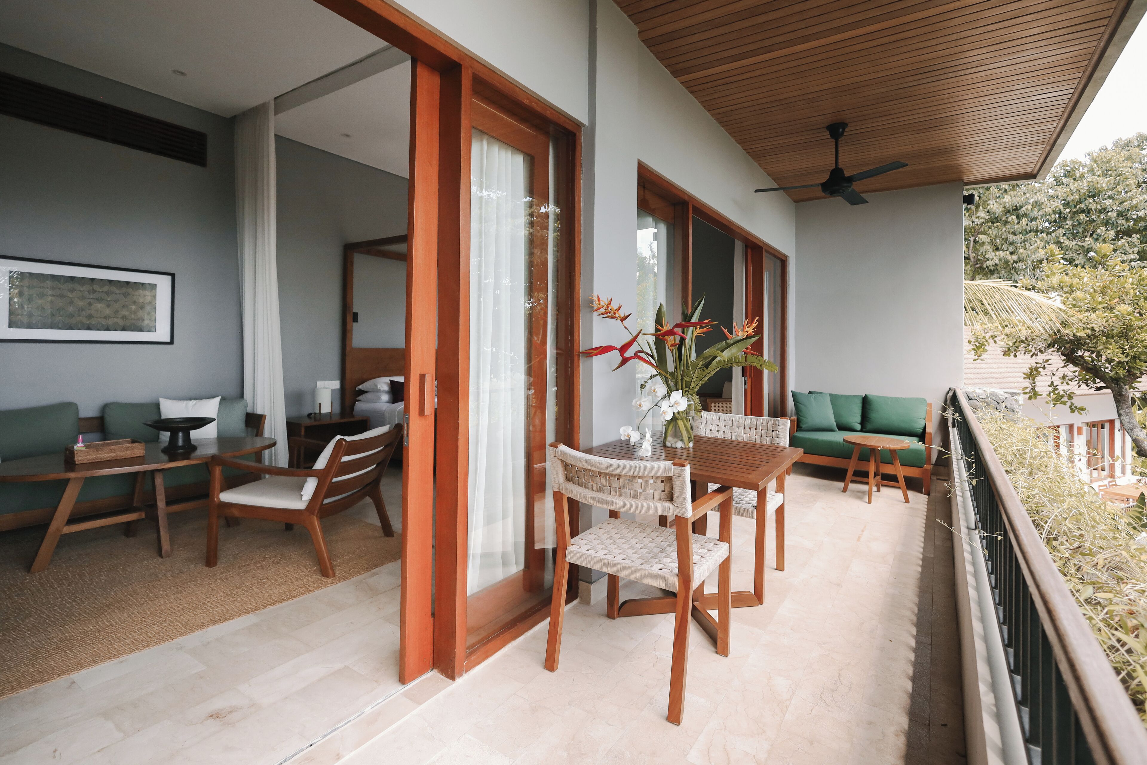 A modern outdoor seating area with wooden furniture, a table with a vase of flowers, and a view of greenery.