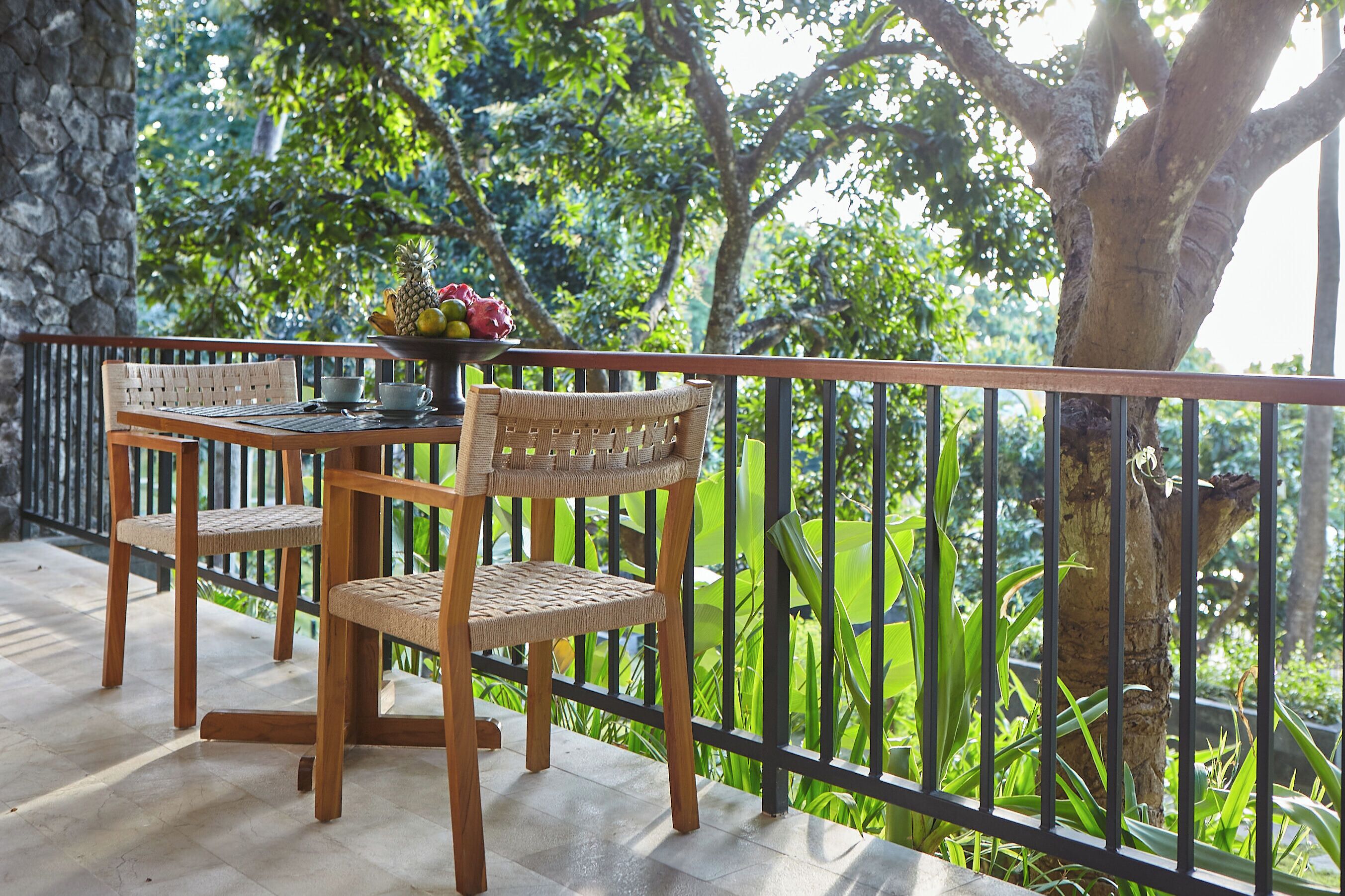 Una mesa y sillas de madera en un balcón con vista a exuberante vegetación y un muro de piedra.