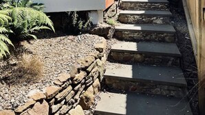 Un escalier en pierre mène à une terrasse en bois, adossée à un mur en briques et dotée d’un espace abrité.