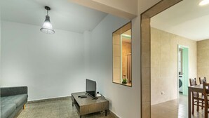 A modern living room with a grey sofa, a coffee table, a TV, and a pendant light. A large mirror reflects a bathroom with a washing machine.