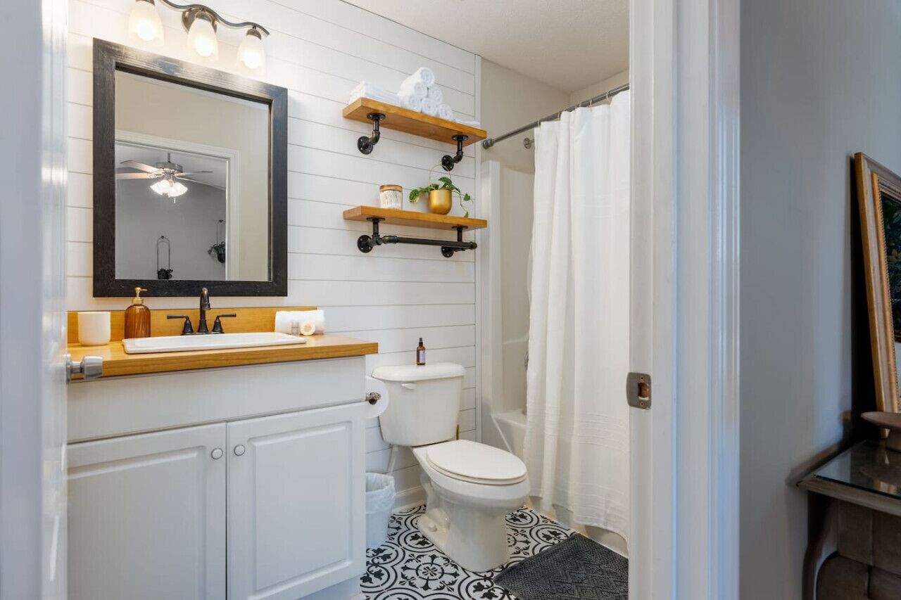 A bathroom with a white sink, a toilet, a shower curtain, and a mirror with lights. There are wooden shelves above the sink and a plant on one of them.