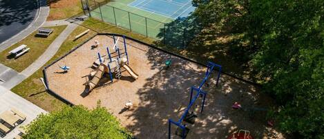 A playground with various play structures, a tennis court, and a surrounding fence.