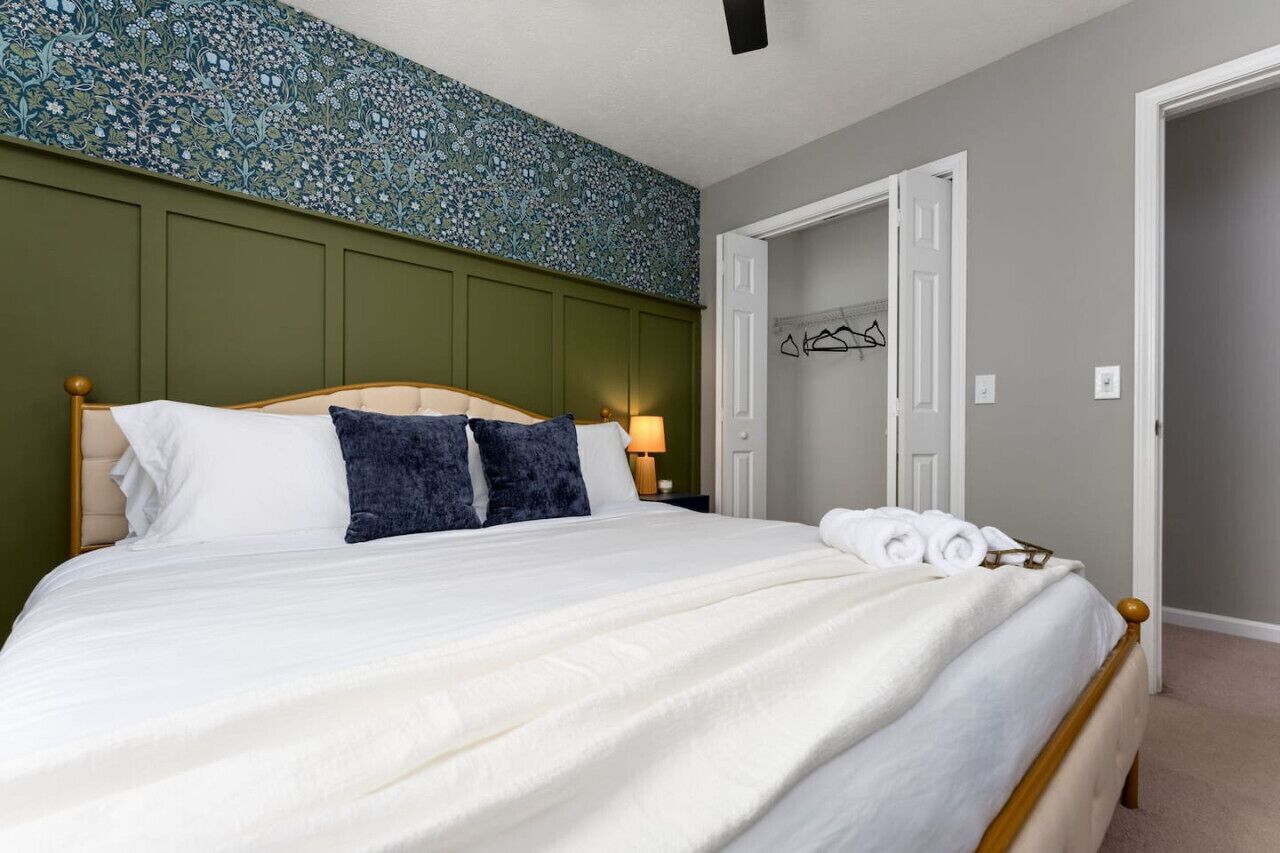 Un lit bien fait, avec des oreillers blancs et bleu foncé, une tête de lit en bois et un mur vert orné d’un motif floral.