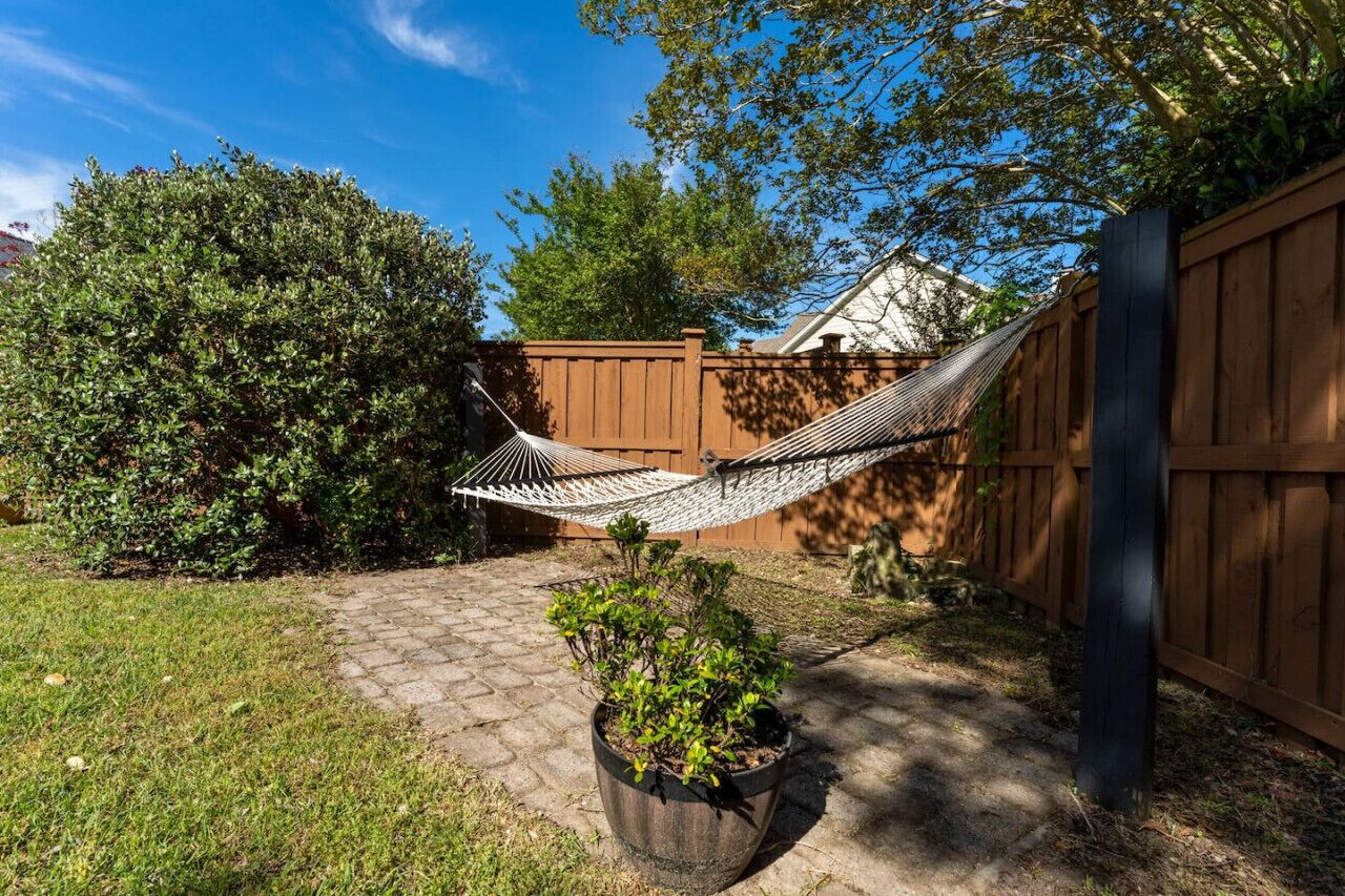 A hammock between a wooden fence and a tree, a potted plant, and a paved pathway in a backyard.