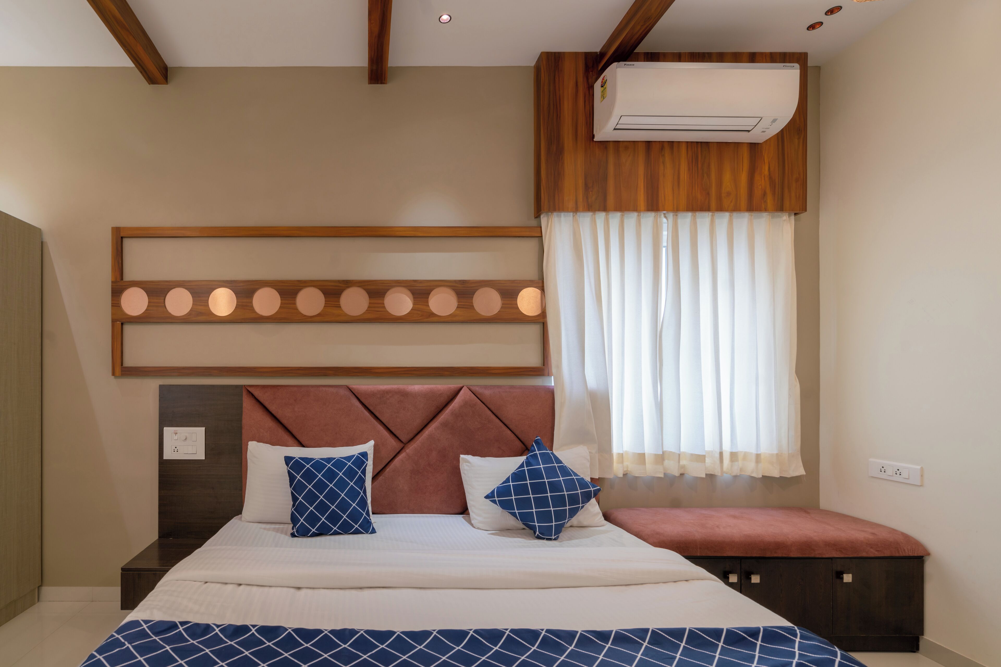 A bedroom with a bed, a headboard, a bedside table, a bench, an air conditioning unit, and a window with curtains.