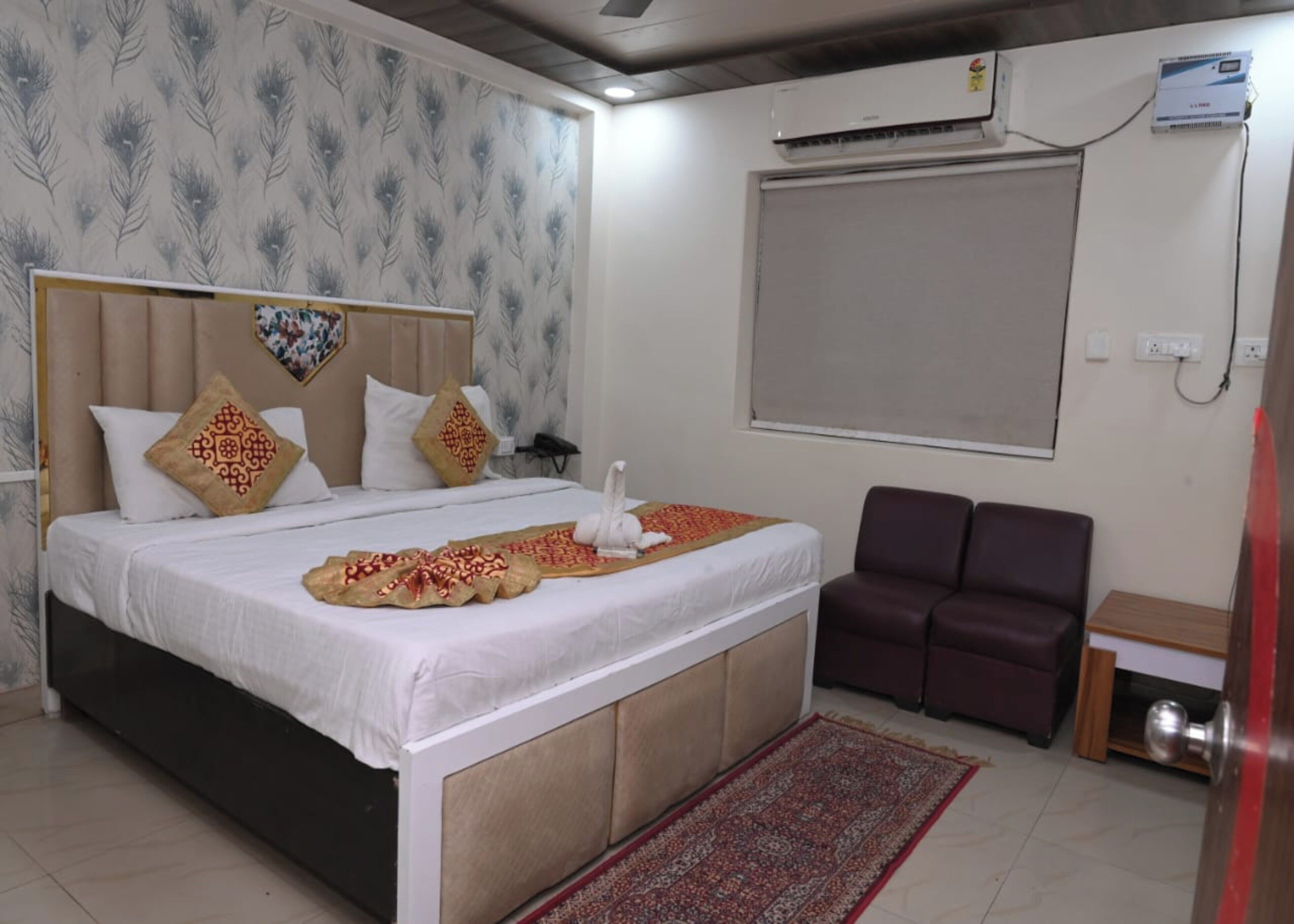 A neatly arranged hotel room with a bed, a small seating area, a wall-mounted air conditioner, and a window with a blind.