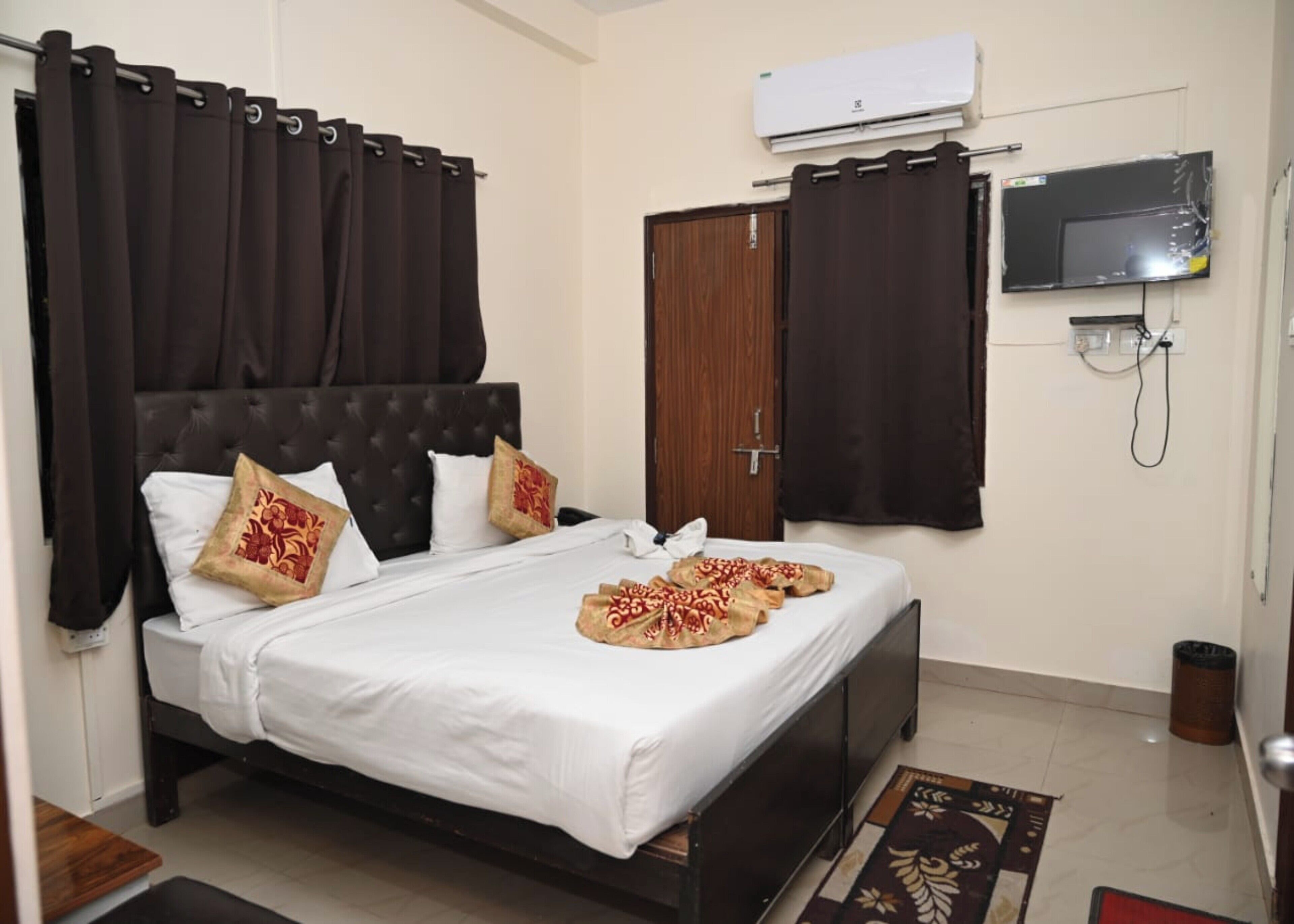 A bedroom with a bed, dark curtains, a wooden door, an air conditioner, a TV, and a trash bin.