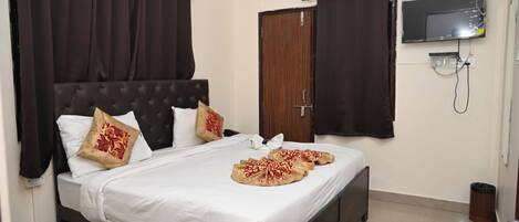 A bedroom with a bed, dark curtains, a wooden door, an air conditioner, a TV, and a trash bin.