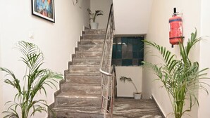 A staircase with marble steps, a metal handrail, and a red carpet. There are potted plants on either side and a fire extinguisher on the wall. - Hotel Aparajita (Varanasi)