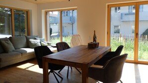 Un moderno salón con una mesa de comedor de madera, sillas negras y un sofá con cojines. Amplias ventanas ofrecen luz natural y vistas al exterior.