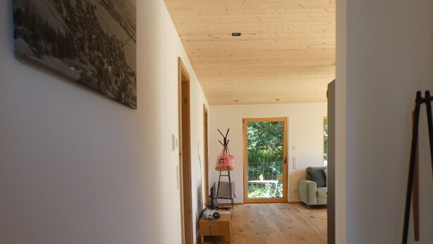 Un pasillo con suelo de madera, paredes blancas y techo con vigas de madera. Se observa un cuadro en la pared izquierda y una puerta que da a otra habitación.