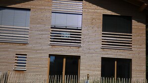 Un edificio moderno con listones horizontales de madera, ventanas amplias y una valla metálica al frente.