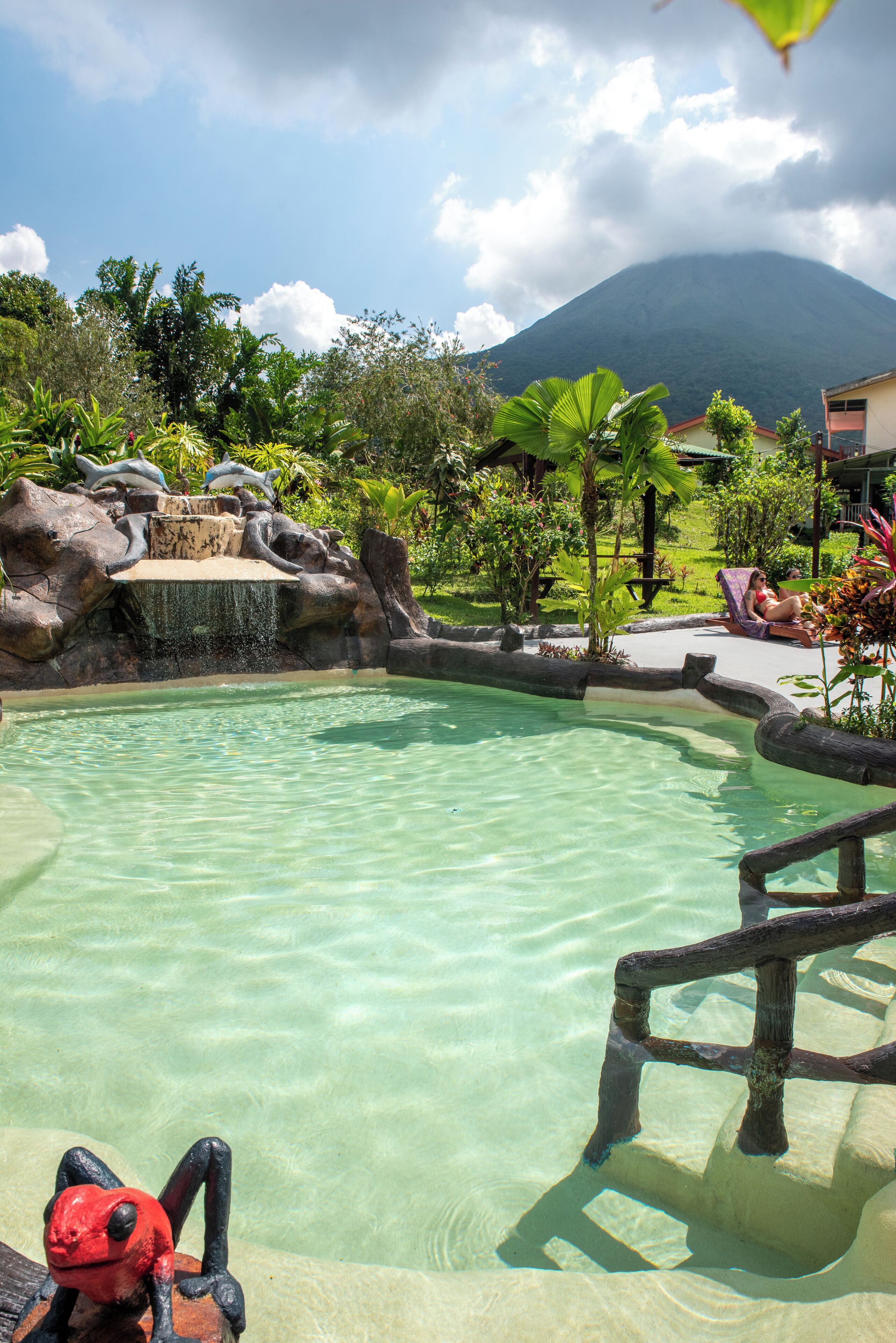 A clear pool with a waterfall feature, surrounded by lush greenery and a mountain in the background.
