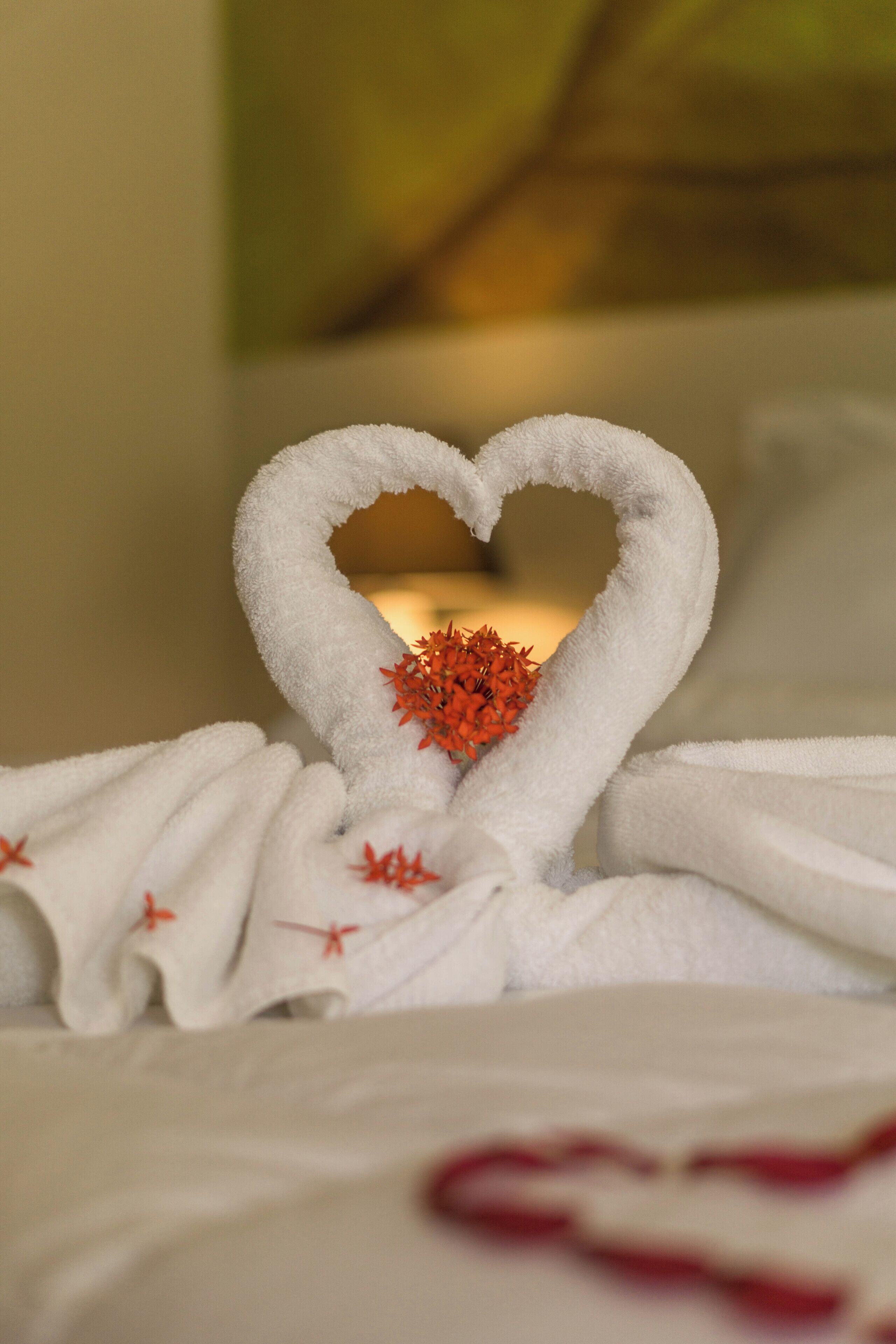 A bed with a heart-shaped towel arrangement and a small red flower centerpiece.