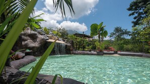A clear pool with a waterfall feature surrounded by tropical vegetation and a distant building.
