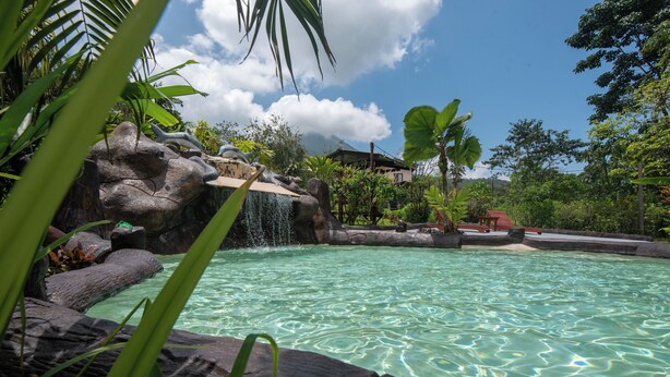 A clear pool with a waterfall feature surrounded by tropical vegetation and a distant building. - Sueno Dorado Hot Springs Hotel (La Fortuna)