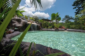 Une piscine aux eaux cristallines, agrémentée d’une cascade, entourée d’une végétation tropicale et avec un bâtiment visible au loin.