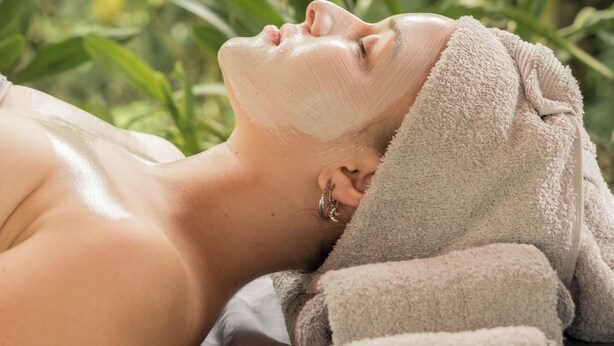 A person lying down with a towel over their head, in a room with a wooden railing and greenery in the background.
