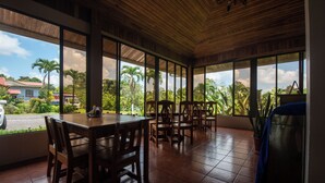 A covered dining area with wooden tables and chairs, large windows offering a view of palm trees and houses. - Sueno Dorado Hot Springs Hotel (La Fortuna)