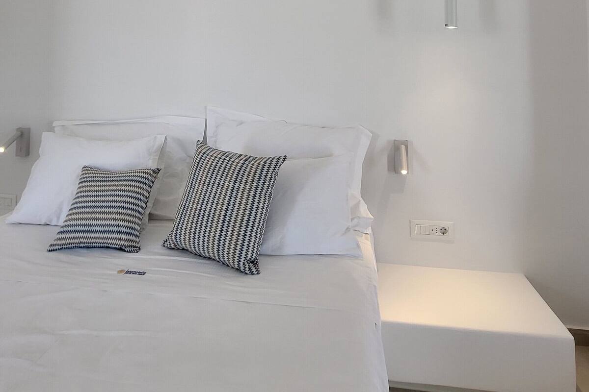 A modern bedroom with a large bed, two patterned pillows, a bedside table, and a wall-mounted light fixture.