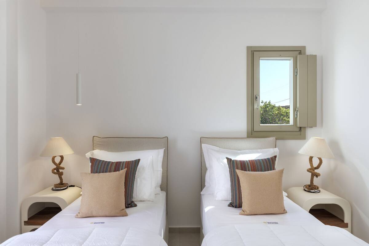 A double room with two beds, each with a nightstand, a lamp, and a window with a view of greenery.