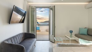 En moderne stue med en grå sofa, en hvid lænestol og et glasbord. Et stort vindue giver udsigt over havet og bjergene.