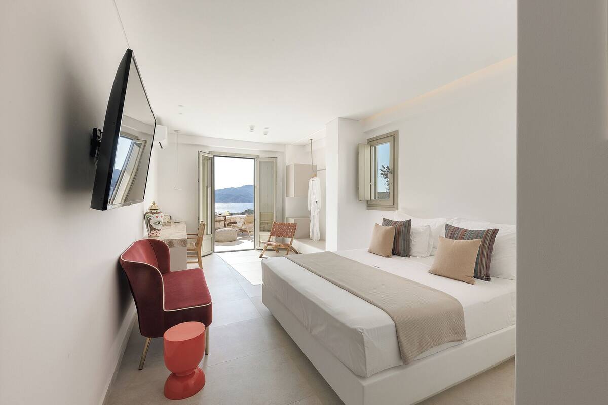 A modern bedroom with a bed, a desk, and a chair. There is a view of the sea through an open door.