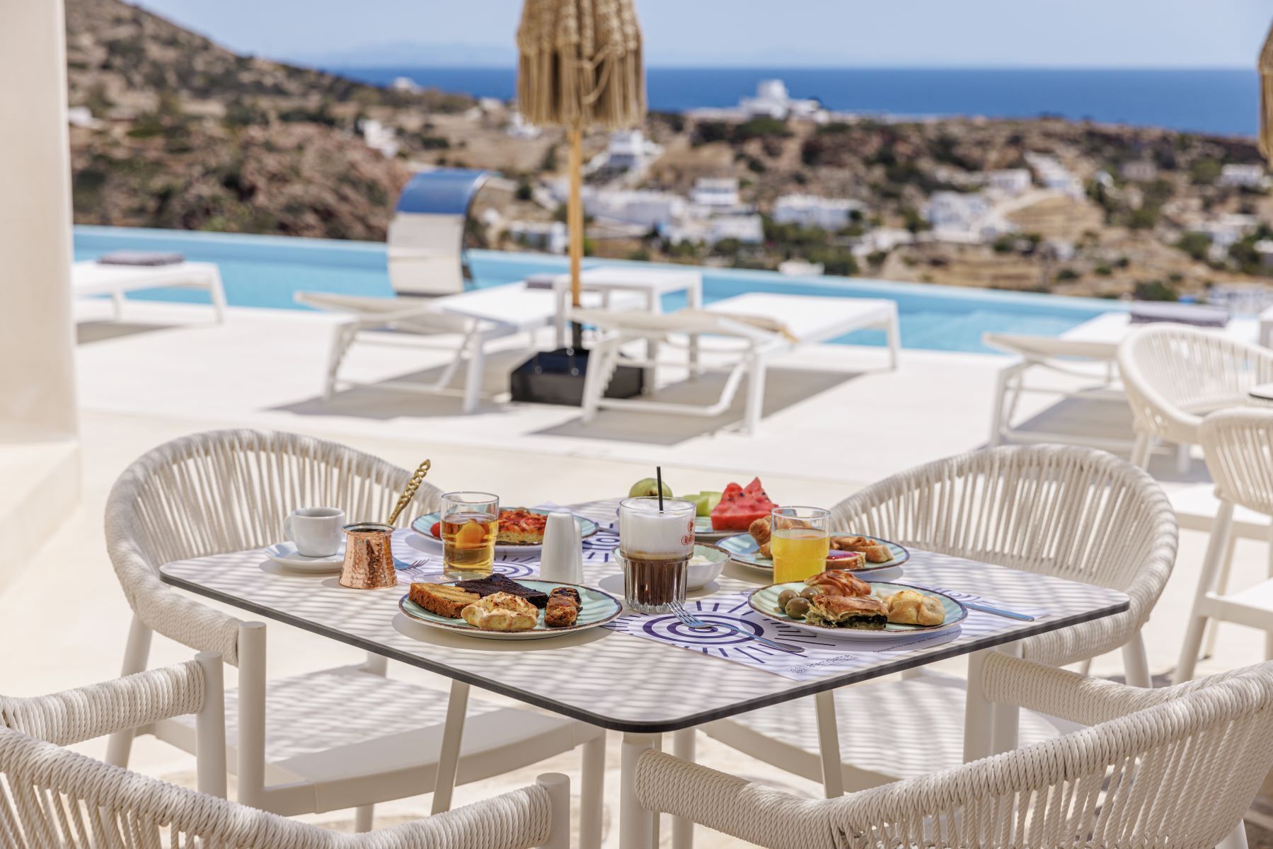 A table set with food and drinks on a terrace overlooking a pool and a coastal view.