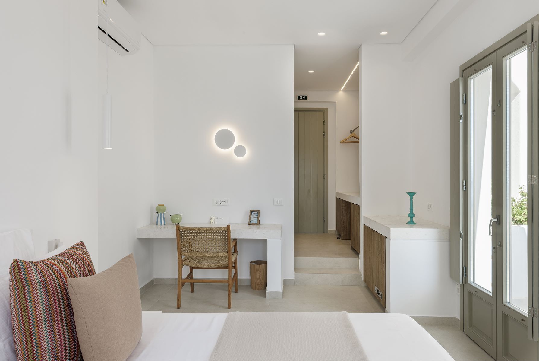 Un dormitorio moderno con cama, escritorio y silla. Hay una puerta que da a un baño y un pasillo.