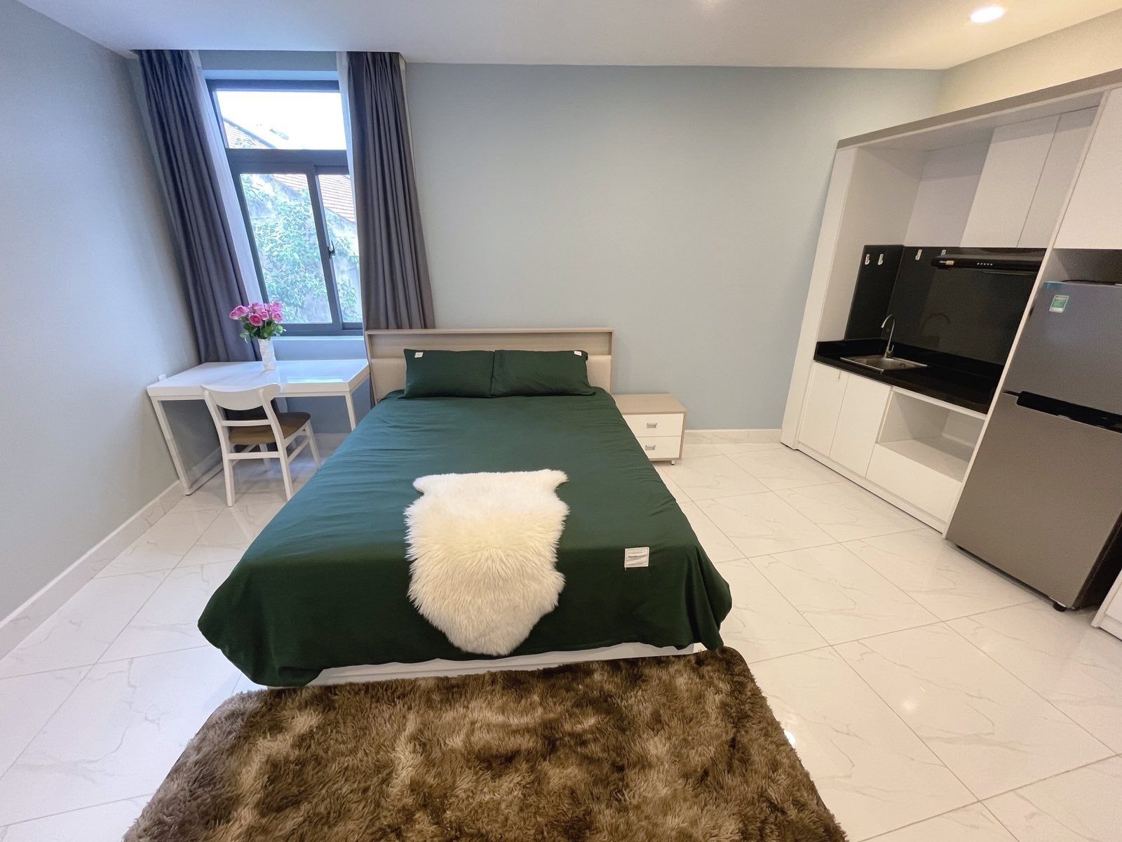 A bedroom with a bed, a desk, a chair, a small table with flowers, a window with curtains, and a kitchenette with a fridge and oven.