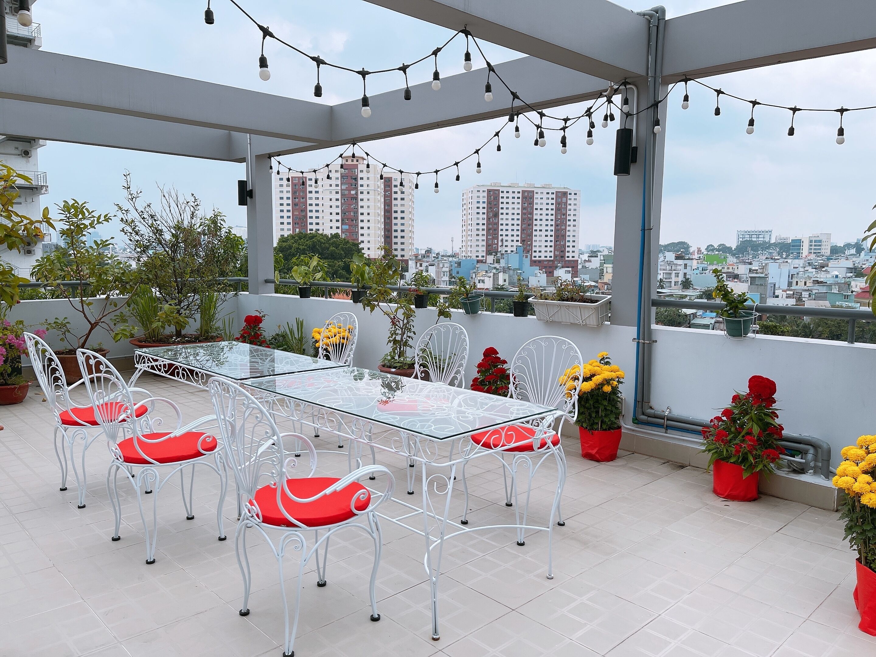 A rooftop terrace with a glass table and white metal chairs, surrounded by potted plants and flowers.