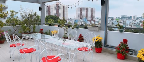 A rooftop terrace with a glass table and white metal chairs, surrounded by potted plants and flowers.