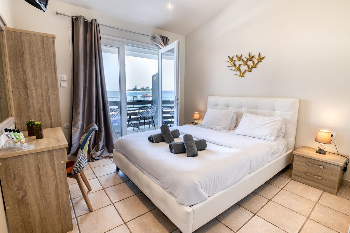 A hotel room with a large bed, a desk with toiletries, a chair, a nightstand with a lamp, and a balcony with a view of the sea.