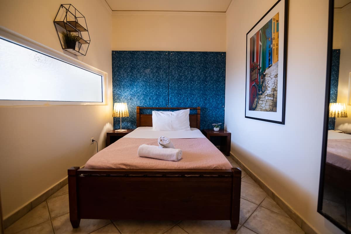 A neatly made bed with a pink bedspread, a white pillow, and a rolled towel on the footboard. There is a nightstand with a lamp and a small potted plant beside the bed. The room has a blue textured wall behind the bed and a framed picture on the wall to the right.