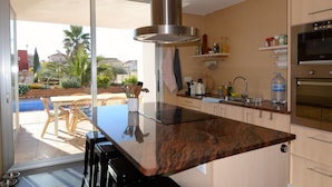 A modern kitchen with a large island, a built-in oven, and a sliding glass door leading to a patio with outdoor furniture. - VILLA PONENT (L'Escala)