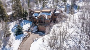 Un grand chalet à plusieurs étages, dont le toit est recouvert de neige, entouré d’arbres et accessible par un chemin dégagé menant à l’entrée.